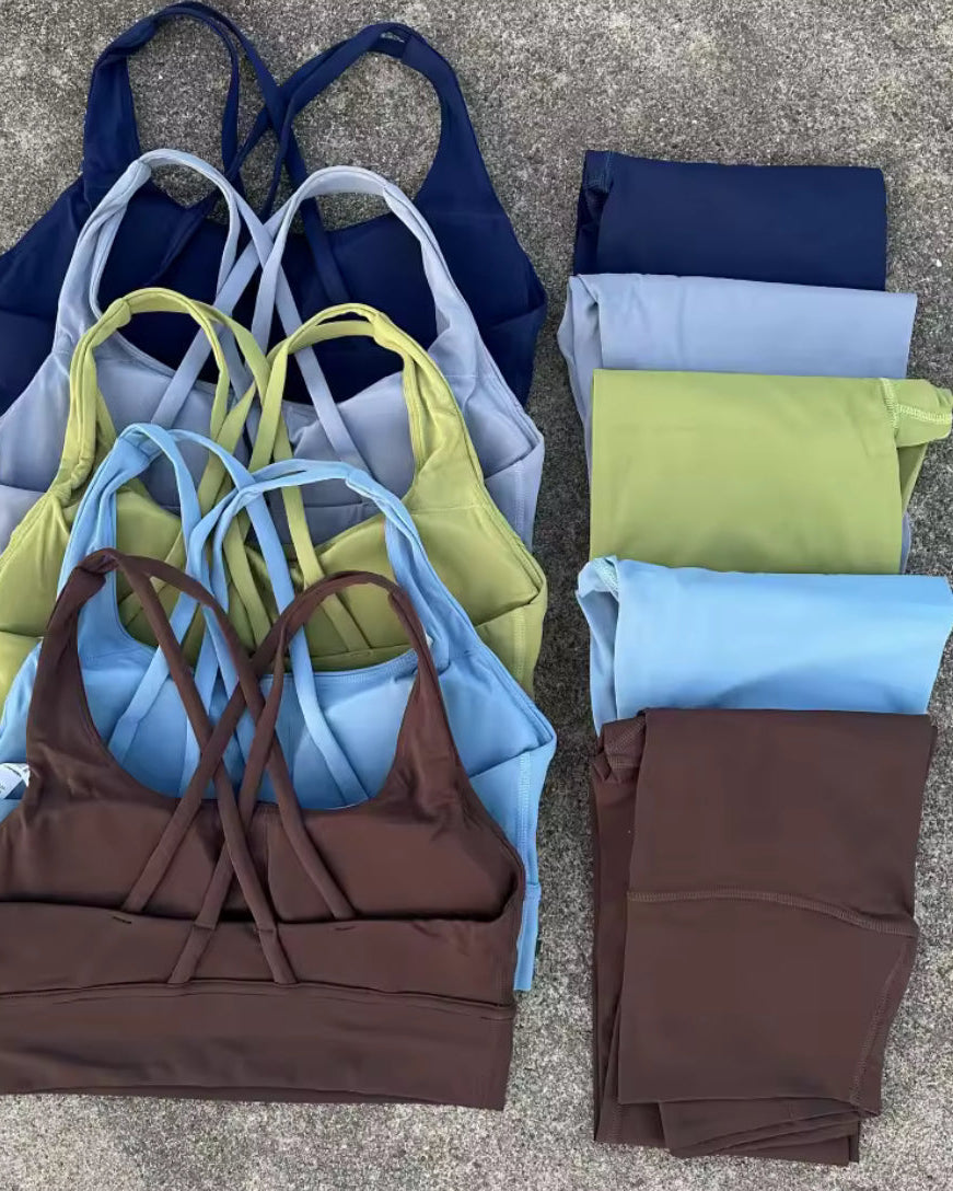 Six pairs of matching activewear sets laid out on a gray concrete surface, featuring sports bras and leggings in navy, light blue, olive green, and brown.