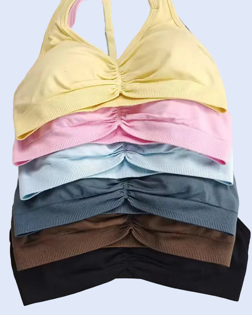 A stack of women's wireless ruched sports bra cups in pastel yellow, pink, light blue, slate blue, brown, and black, arranged on a white surface with decorative props.