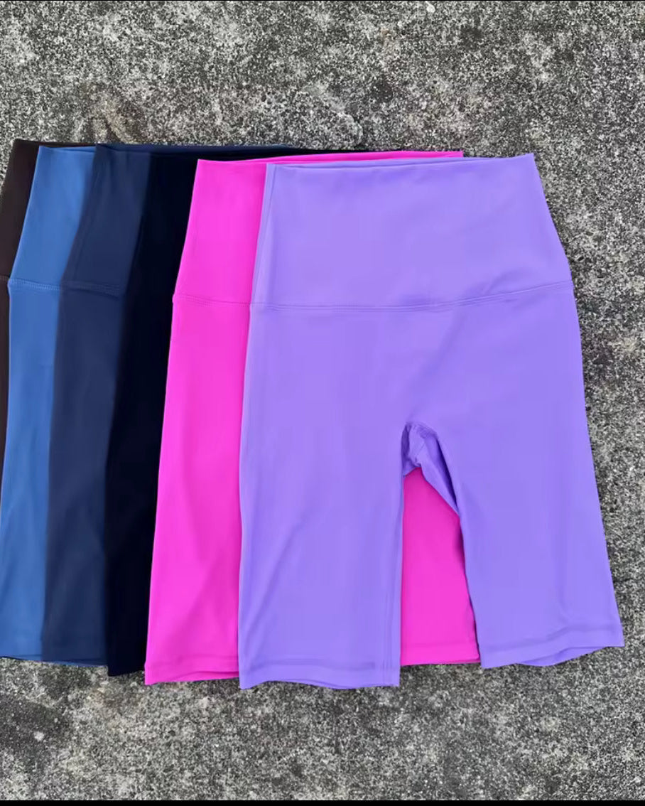 Three pairs of high-waisted athletic biker shorts in purple, pink, and navy blue, laid flat on a textured gray concrete surface.