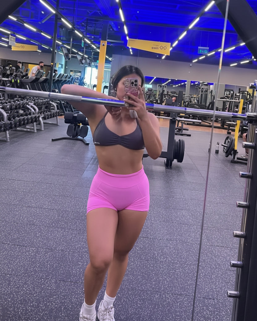 Person taking a mirror selfie in a gym wearing a sports bra and pink shorts.