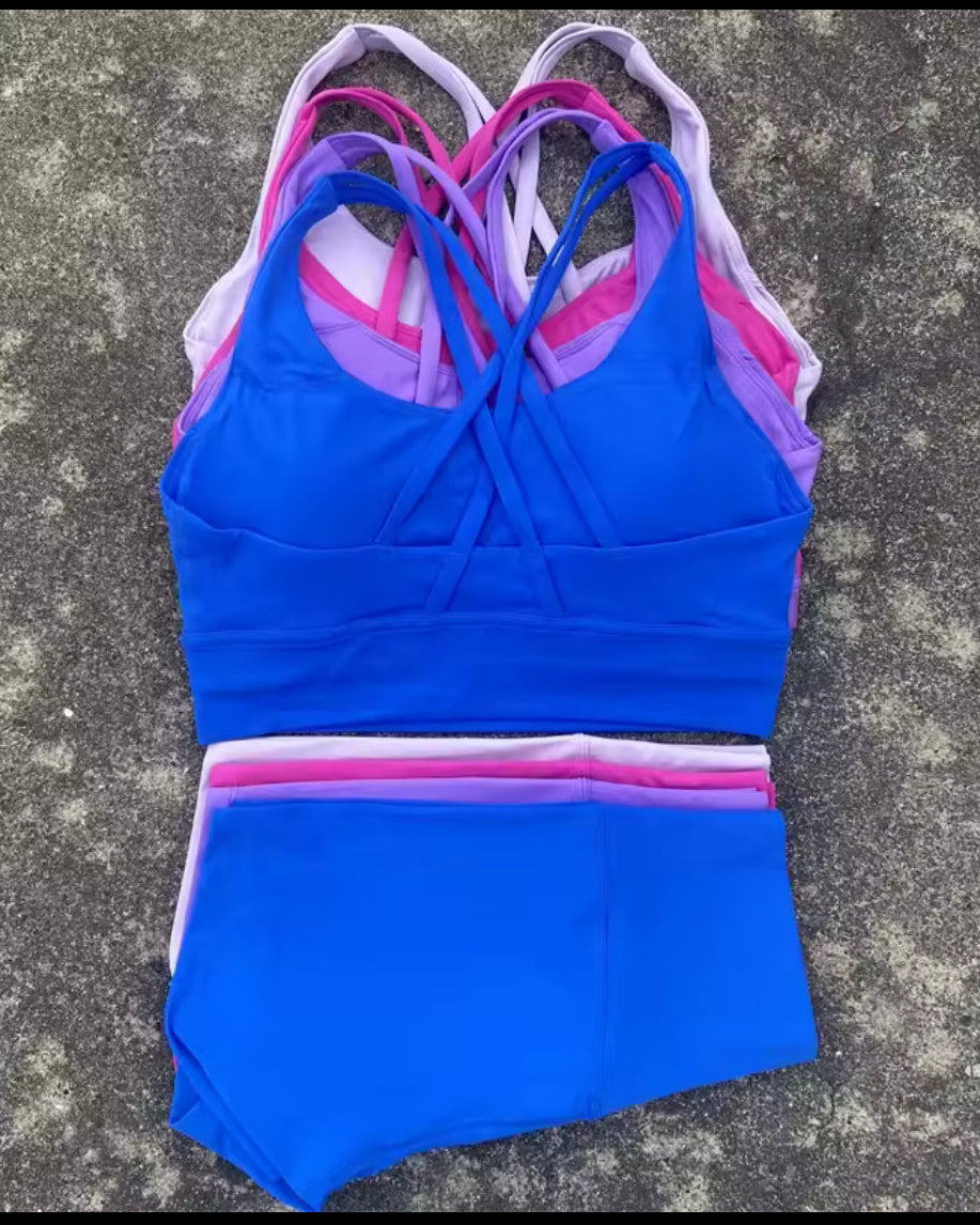 A two-piece athletic swimsuit set laid flat on a gray concrete surface, featuring a bright blue sports bra top with layered pink, purple, and white straps, and matching high-waisted blue bottoms with a striped waistband.