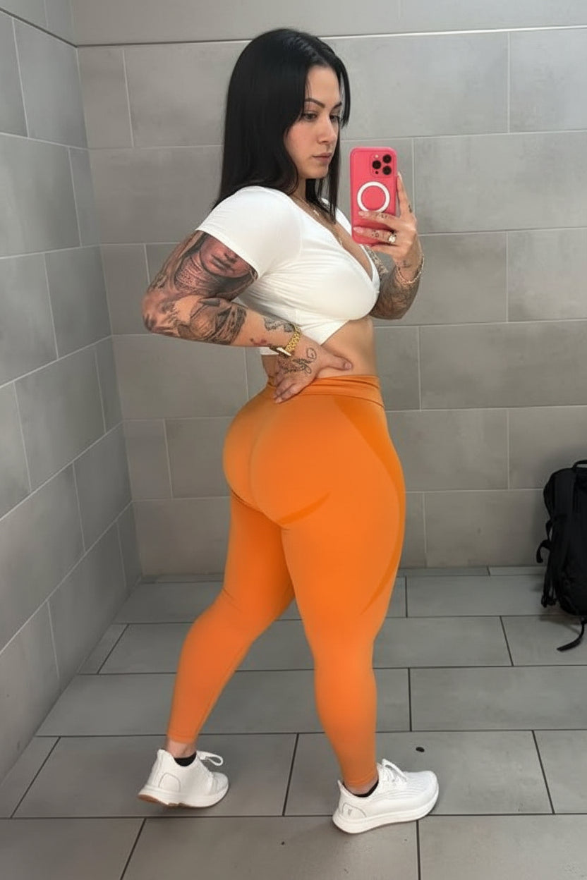 Woman taking a mirror selfie wearing a white top and orange leggings in a tiled room.