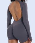 A woman modeling a dark gray long-sleeve bodysuit with a dramatic open back, shown from a rear three-quarter angle against a light gray studio background.
