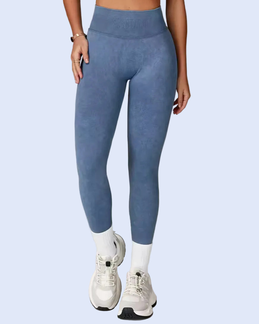 A model wearing a pair of high-waisted, ankle-length athletic leggings in a dusty medium blue color, styled with white socks and white sneakers against a light gray studio background.
