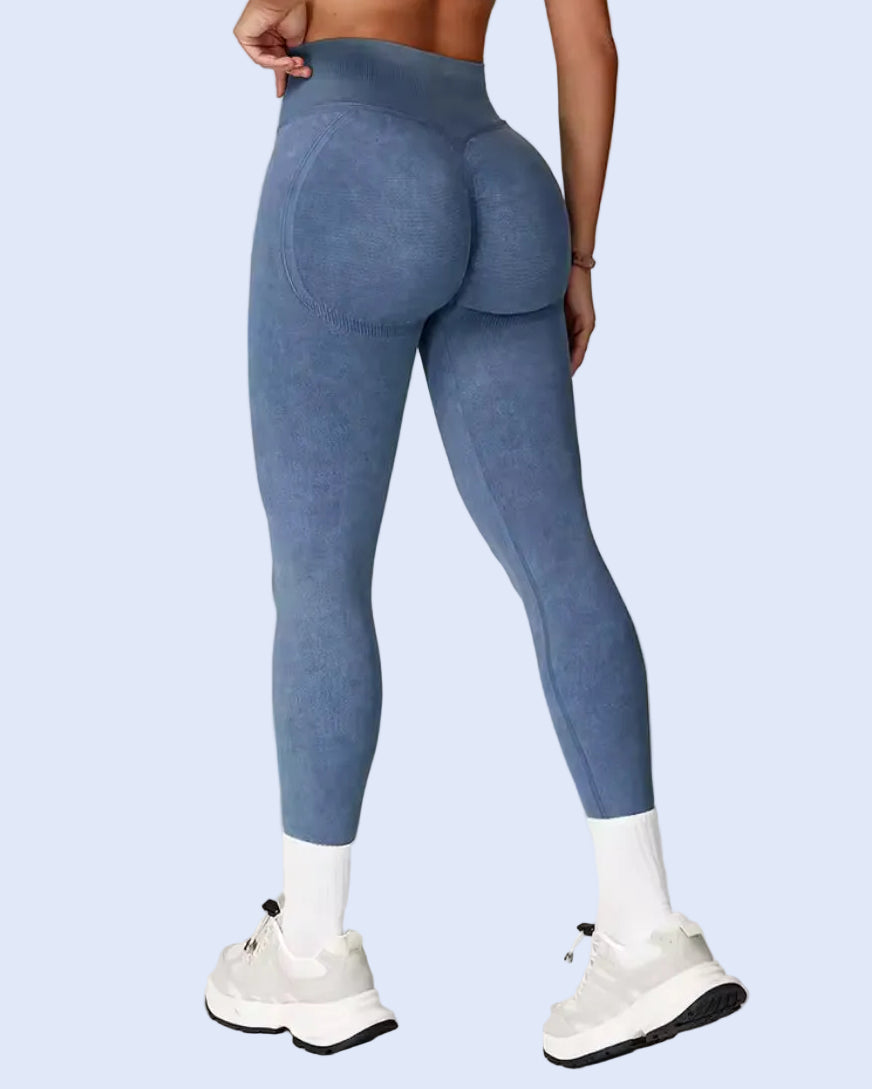 A person shown from the back wearing high-waisted blue leggings with a textured, suede-like finish, white socks, and white sneakers against a light gray studio background.