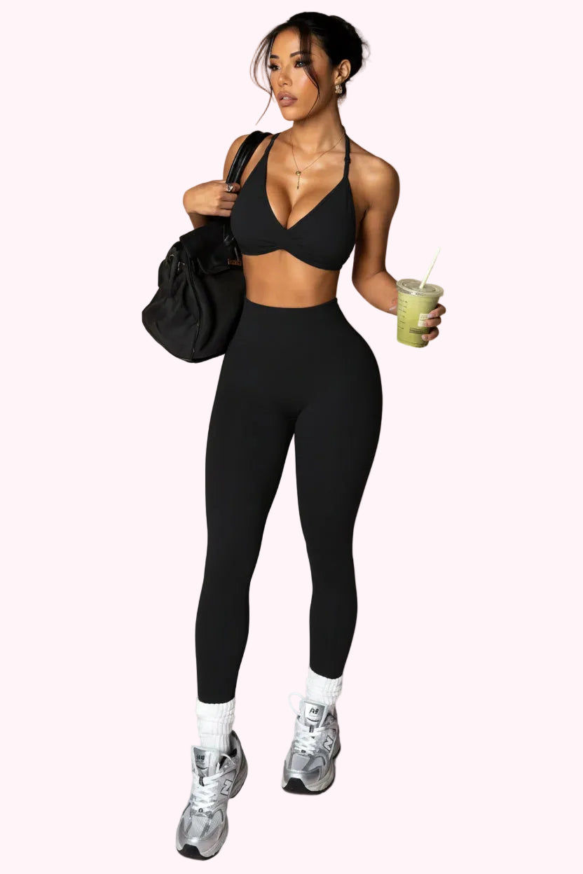 Woman in black athletic wear holding a green drink and black bag on a plain background