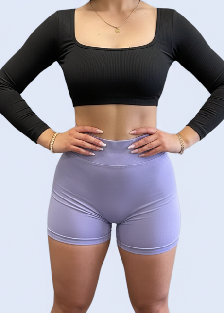 Woman taking a mirror selfie in a gym wearing a black long-sleeve top and light purple shorts.