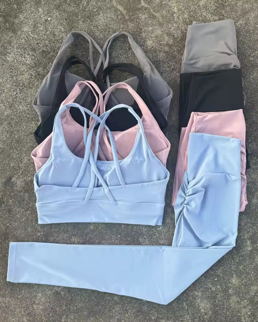 A set of athletic wear laid flat on a gray concrete surface, including a light blue sports bra with crisscross straps, matching light blue leggings, a stack of folded leggings in gray, black, and pink, and a separate light blue leggings section shown separately.