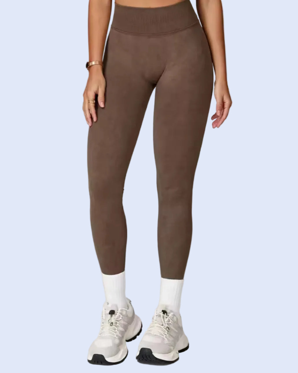 A model wearing high-waisted, ankle-length athletic leggings in a solid taupe-brown color, paired with white socks and white sneakers, against a light gray studio background.
