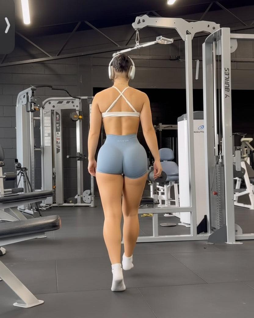 A woman from behind wearing a white criss-cross sports bra and light blue high-waisted leggings, walking through a gym.