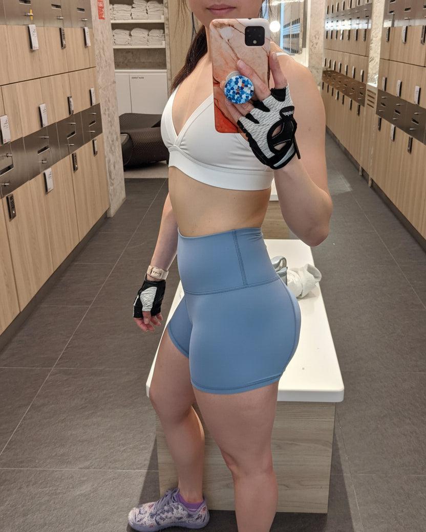 Person taking a mirror selfie in a locker room wearing a white sports bra and blue shorts.