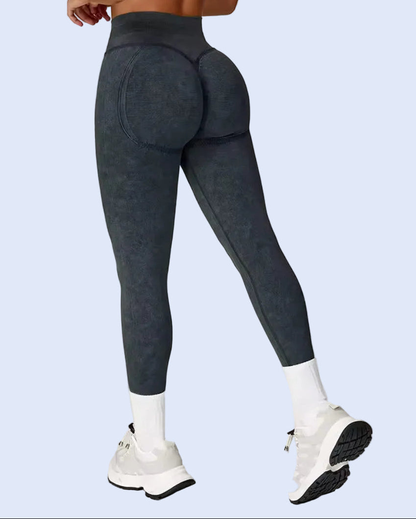 Rear three-quarter view of a model wearing dark gray high-waisted athletic leggings with a textured finish, paired with white socks and white sneakers, against a light gray studio background.