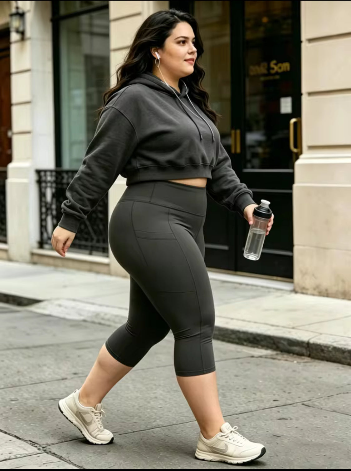 A woman walking on a city sidewalk wearing a dark gray cropped hoodie and matching high-waisted capri leggings, holding a clear water bottle.