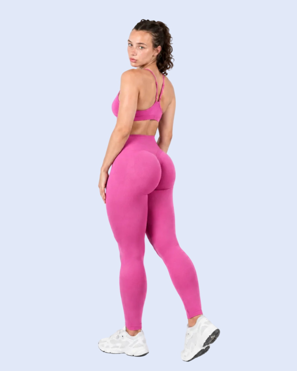 A woman modeling a bright pink activewear set consisting of a sports bra and high-waisted leggings, paired with white sneakers, against a light blue studio background.
