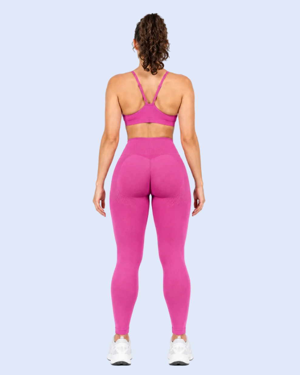 A woman viewed from the back wearing a bright pink sports bra and matching high-waisted leggings, standing against a light blue studio background.