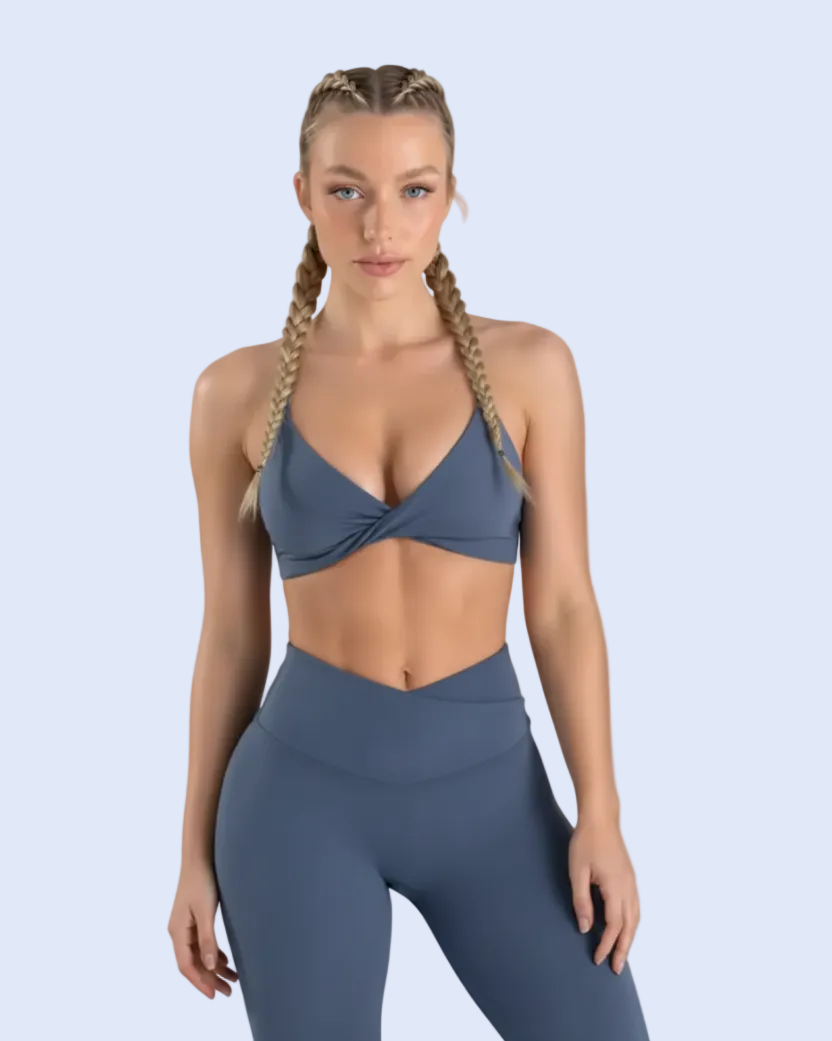 A blonde model wearing a matching slate blue activewear set, featuring a crossover halter sports bra and high-waisted leggings, posed against a dark slate blue studio background.