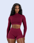 A woman modeling a matching burgundy activewear set consisting of a long-sleeve mock-neck crop top and high-waisted shorts, set against a solid gray studio background.