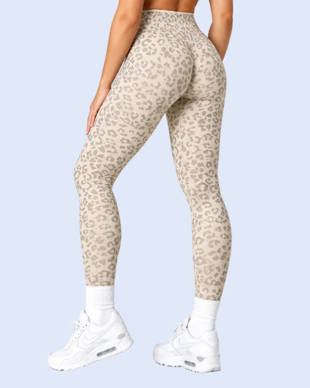 Rear view of a person wearing high-waisted leopard-print leggings in beige and taupe, paired with white socks and white sneakers, against a light blue studio background.