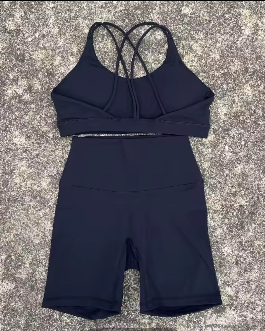 A two-piece athletic set consisting of a dark navy sports bra with a crisscross back strap and matching high-waisted biker shorts, laid flat on a textured gray surface.