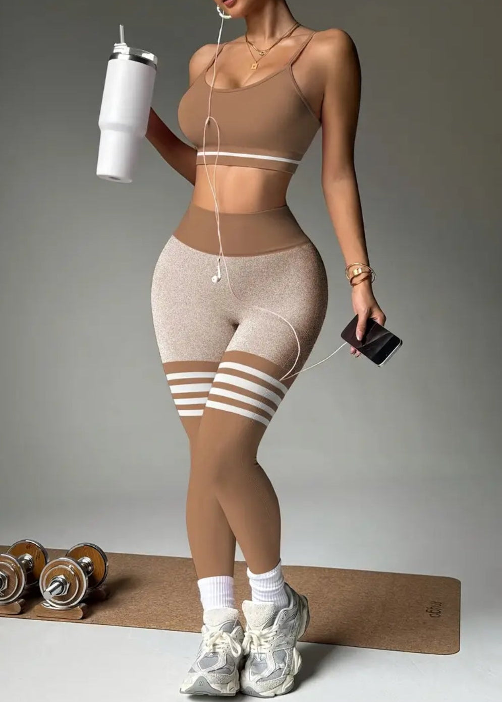 Person wearing a beige athletic outfit with striped leggings, holding a water bottle and phone, on a gray background.