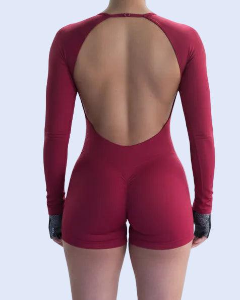 A woman in a gym wearing a form-fitting, long-sleeve, open-back athletic bodysuit in a deep magenta-red color. The photo is taken from a selfie perspective, showing both the back of the model in the mirror reflection and part of her face and shoulder in the foreground.
