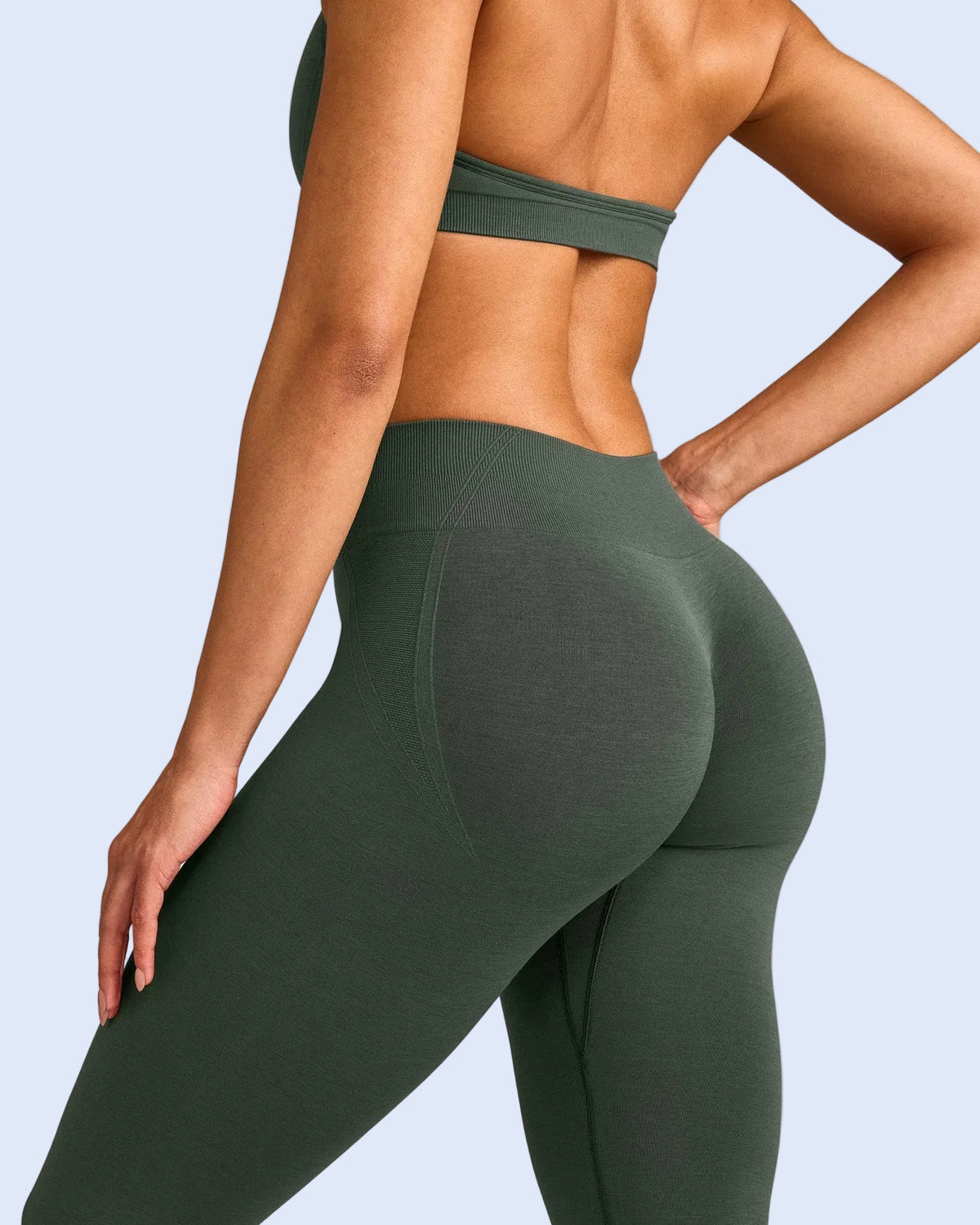 A close-up back and side view of a woman wearing high-waisted, full-length athletic leggings in a muted olive green color, paired with a matching green sports bra, against a solid light gray background.