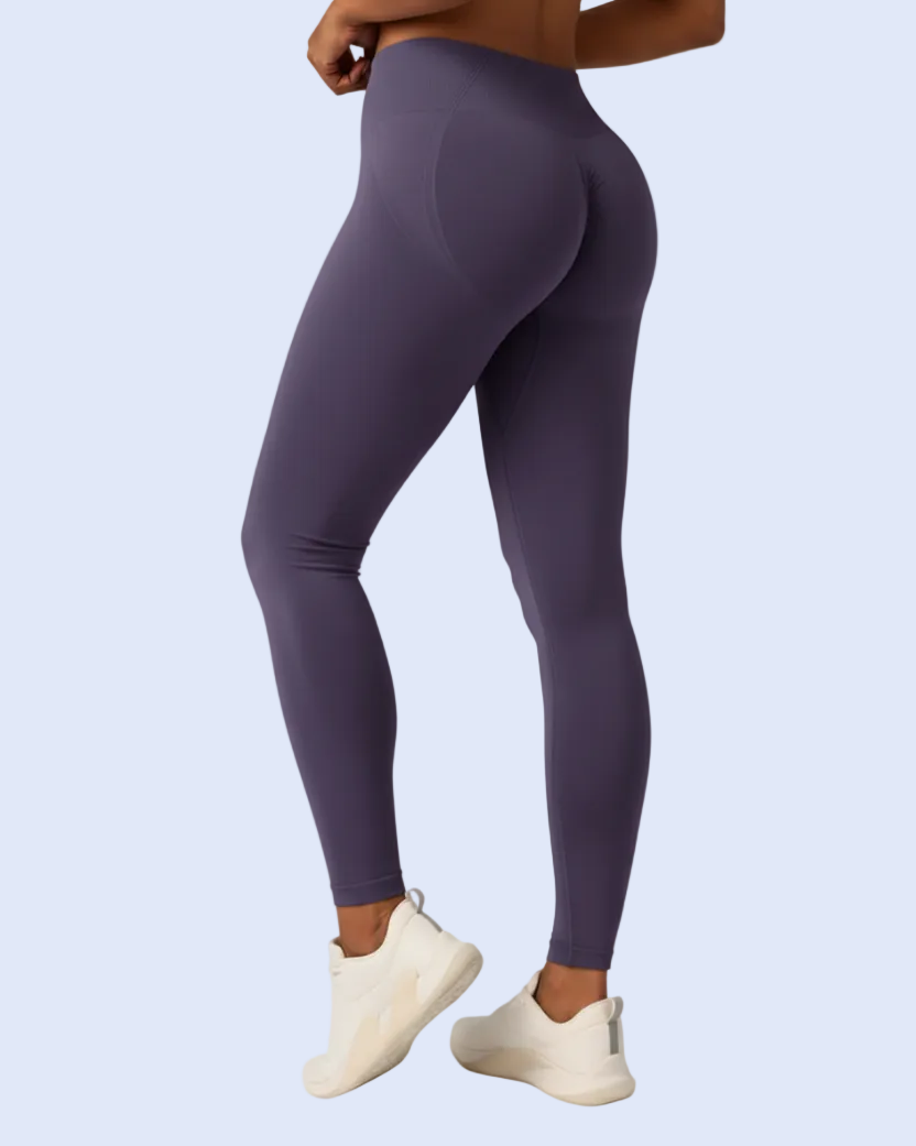 A model wearing high-waisted, form-fitting leggings in a muted purple color, paired with off-white sneakers, shown from a rear three-quarter angle against a light blue background.
