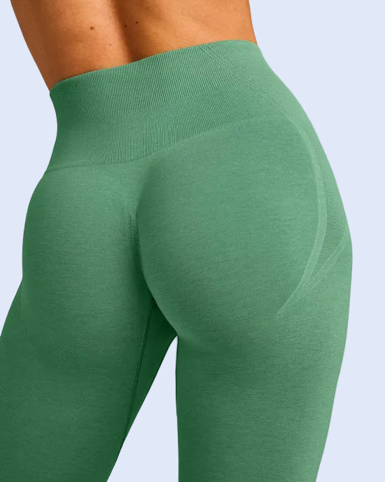 Close-up of a model wearing high-waisted green leggings, showing the ribbed waistband and side hip contour from a rear three-quarter angle against a light gray studio background.