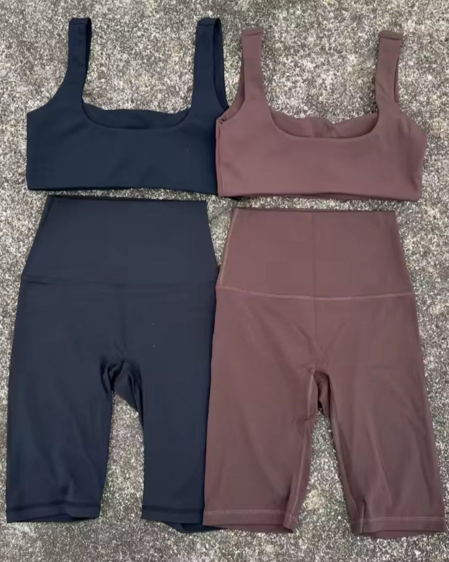 Two matching activewear sets laid flat on a textured gray surface, each consisting of a scoop-neck sports bra and high-waisted biker shorts. The top-left set is dark navy blue, and the top-right set is dusty mauve-brown.