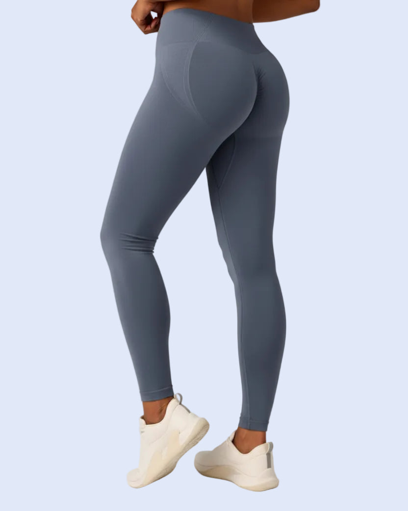 A model wearing slate blue-gray high-waisted leggings with a small white logo near the ankle, paired with beige sneakers, shown from a rear three-quarter angle against a light gray studio background.