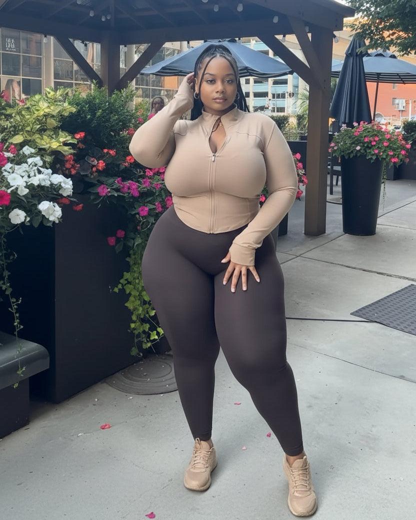 A plus-size woman posing outdoors under a wooden patio canopy, wearing a beige long-sleeve zip-up bodysuit with a keyhole neckline and dark brown leggings.