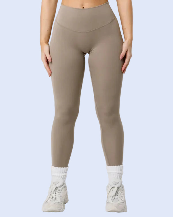 A model from the waist down wearing high-waisted, seamless athletic leggings in a taupe color, paired with white socks and light gray sneakers, against a light gray studio background.