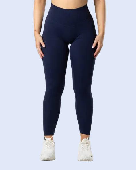 A woman modeling a matching navy blue activewear set consisting of a sports bra and high-waisted leggings, paired with white sneakers, against a light gray studio background.