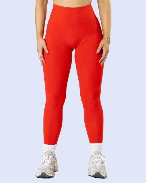A woman modeling a bright red two-piece activewear set consisting of a ruched scoop-neck crop top and high-waisted leggings, paired with white socks and gray sneakers against a plain white studio background.