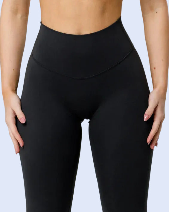 A close-up of a model wearing black high-waisted leggings and a matching black sports bra against a light gray studio background.