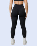 A woman shown from the back wearing a black sports bra and high-waisted black leggings, standing against a light gray studio background.