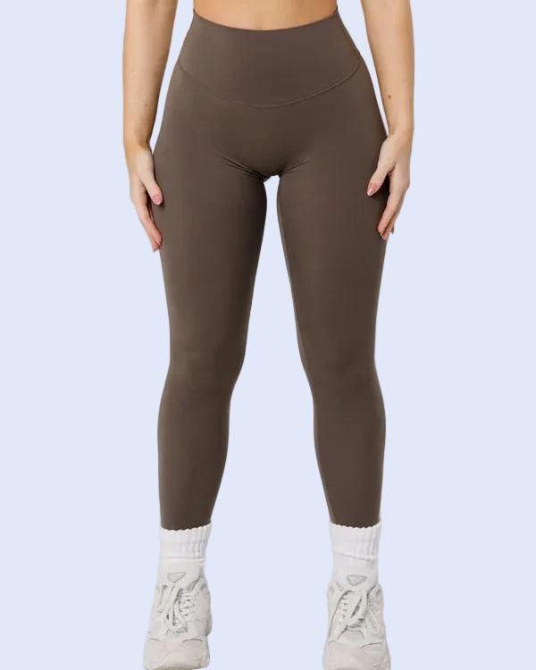 A model wearing high-waisted, ankle-length leggings in a solid taupe-brown color, paired with white socks and white sneakers, against a light gray studio background.