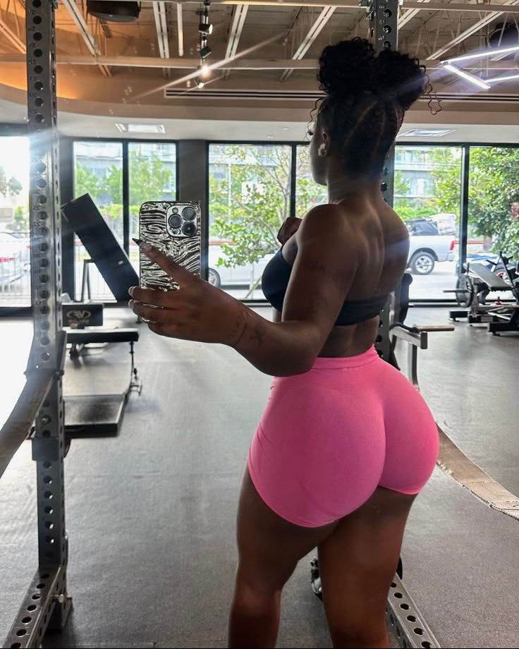 Person taking a mirror selfie in a gym wearing pink shorts and a sports bra.