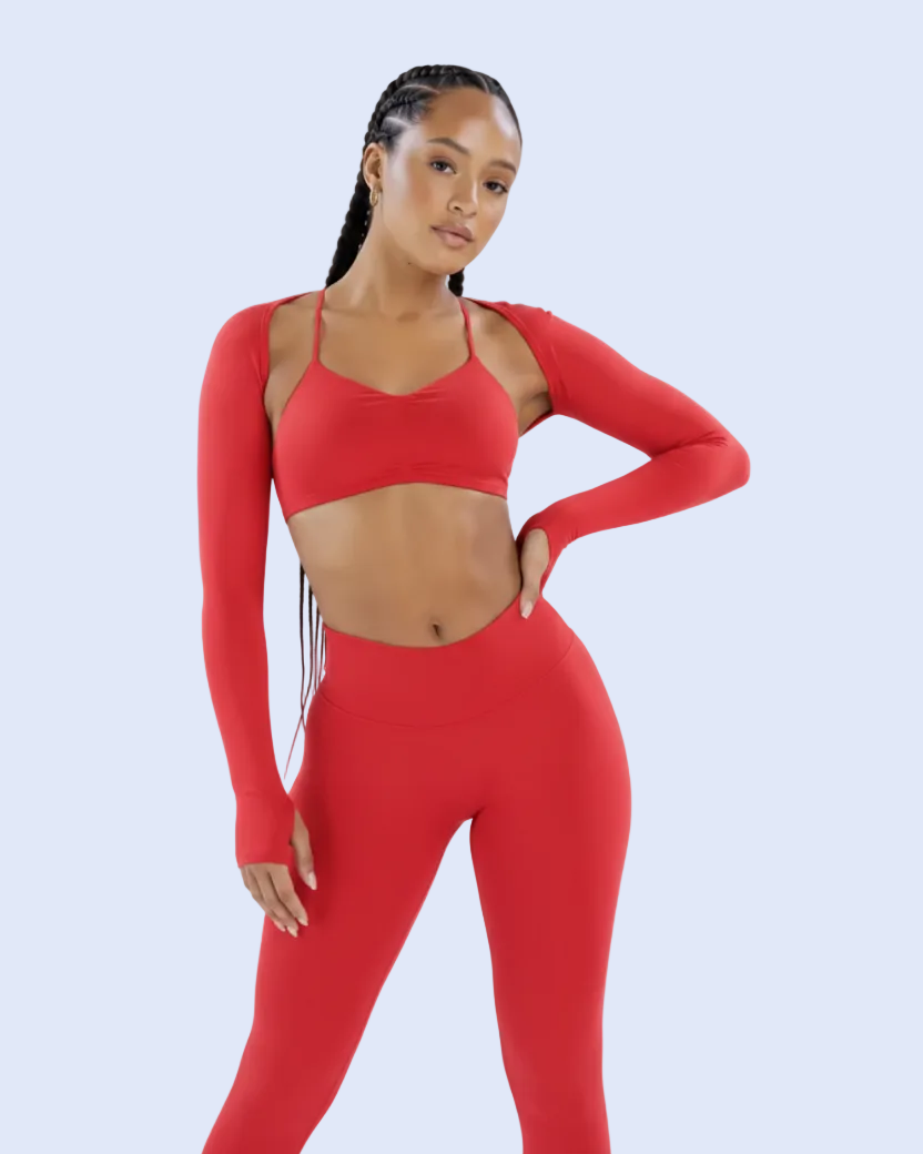 A woman modeling a matching red activewear set consisting of a long-sleeve sports bra and high-waisted leggings, posed against a solid light blue-gray studio background.