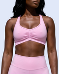 A woman wearing a light pink ruched sports bra and matching high-waisted leggings, standing outdoors against a modern white building and blue sky.