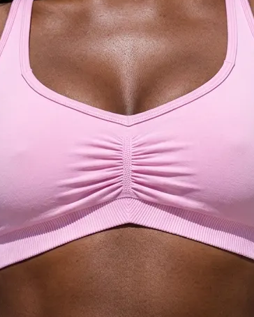 A close-up of a pink sports bra with a scoop neckline and gathered ruching at the center bust, shown on a model.