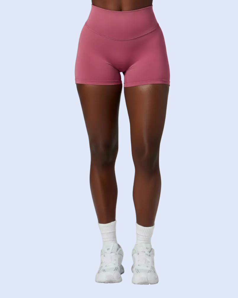A model wearing a matching dusty rose pink activewear set consisting of a sports bra and high-waisted shorts, paired with white socks and sneakers, against a light blue background.