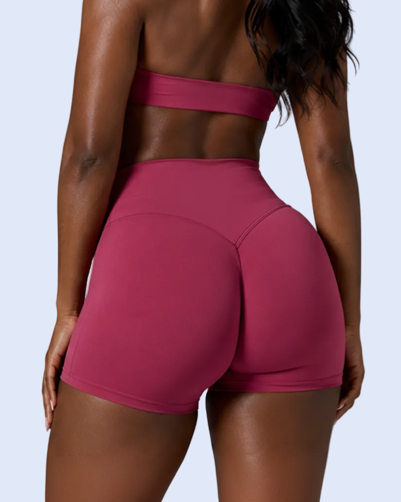 Rear view of a woman wearing magenta athletic shorts and a matching bandeau top with the brand name ACTIVEWEAR BOX, set against a solid gray studio background.