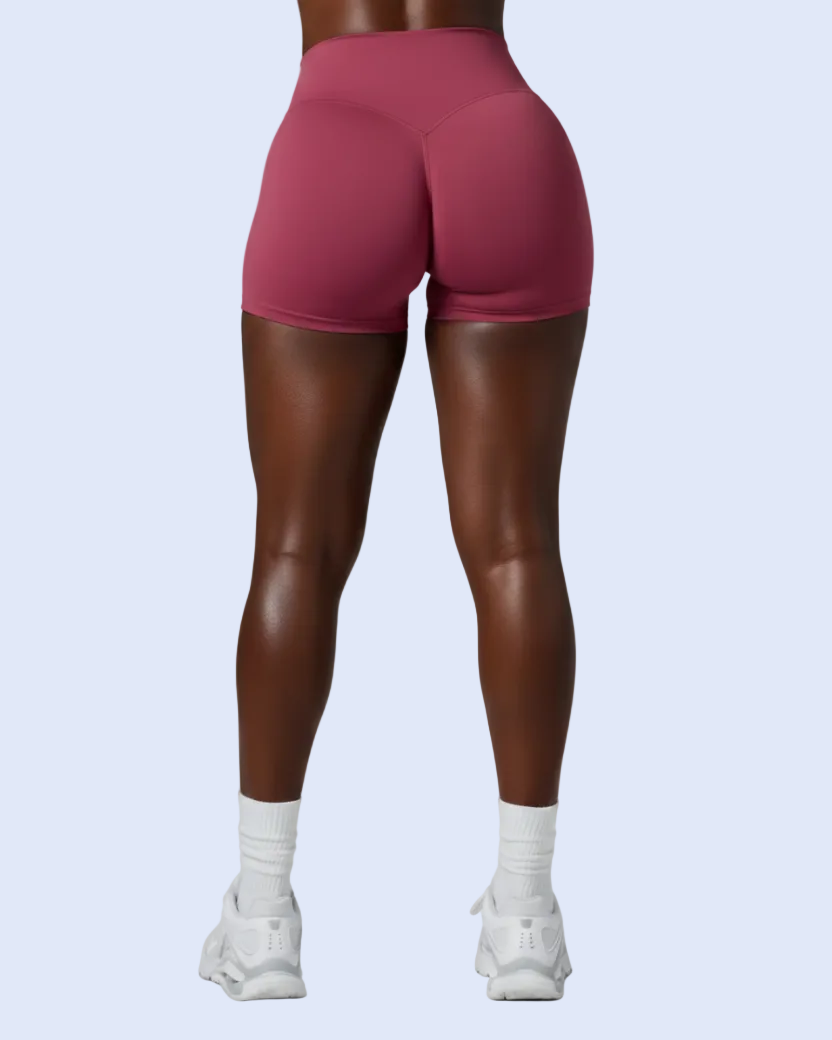 Rear view of a model wearing high-waisted, form-fitting athletic shorts in a dusty rose pink color, paired with white socks and white sneakers, against a light blue background.