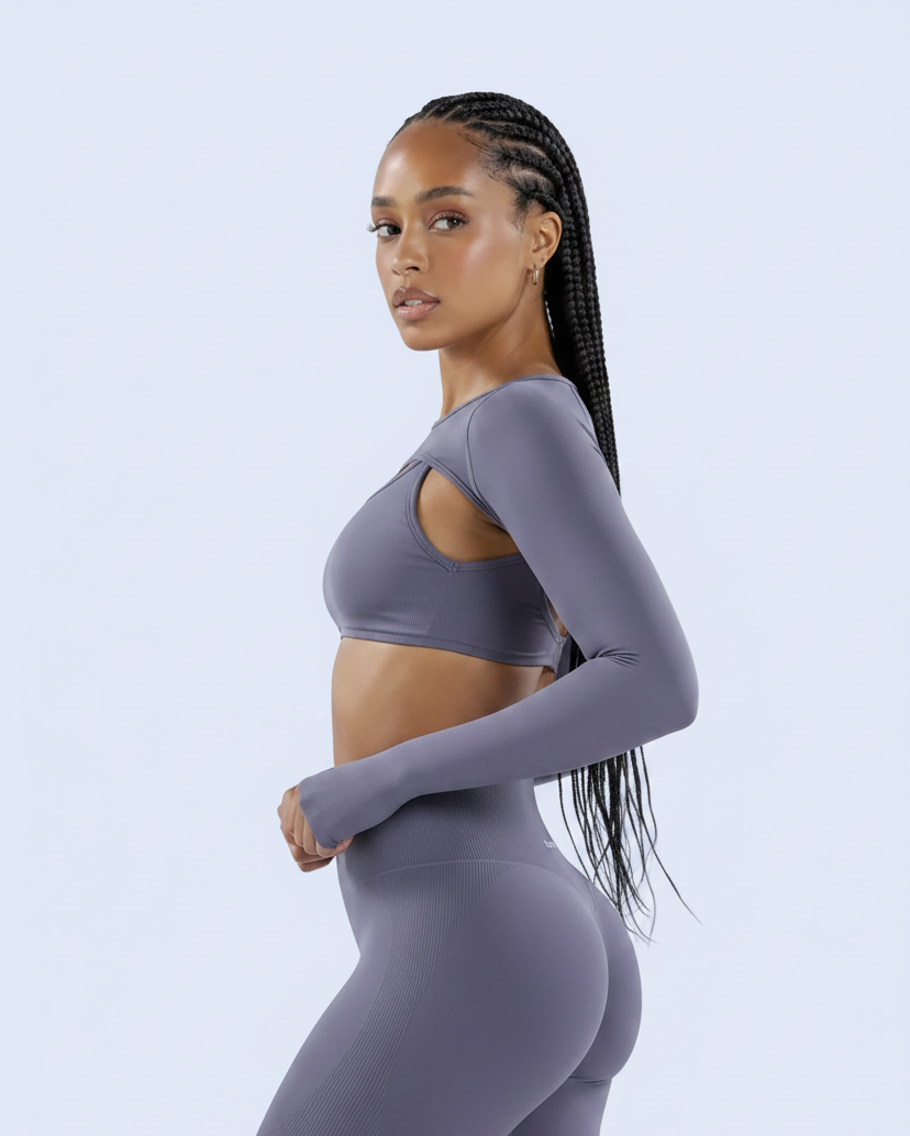 A woman modeling a matching athletic set consisting of a long-sleeve sports bra and high-waisted biker shorts in a dusty purple color, set against a light blue studio background.