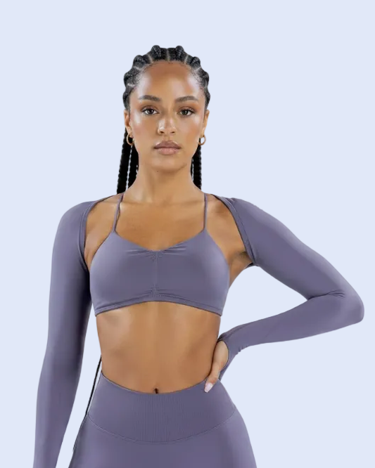 A woman modeling a matching athletic set consisting of a long-sleeve sports bra and high-waisted leggings in a dusty purple color, set against a light blue studio background.