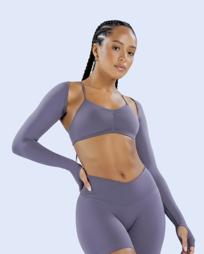 A woman modeling a matching athletic set consisting of a long-sleeve sports bra and high-waisted leggings in a dusty purple color, set against a light blue studio background.