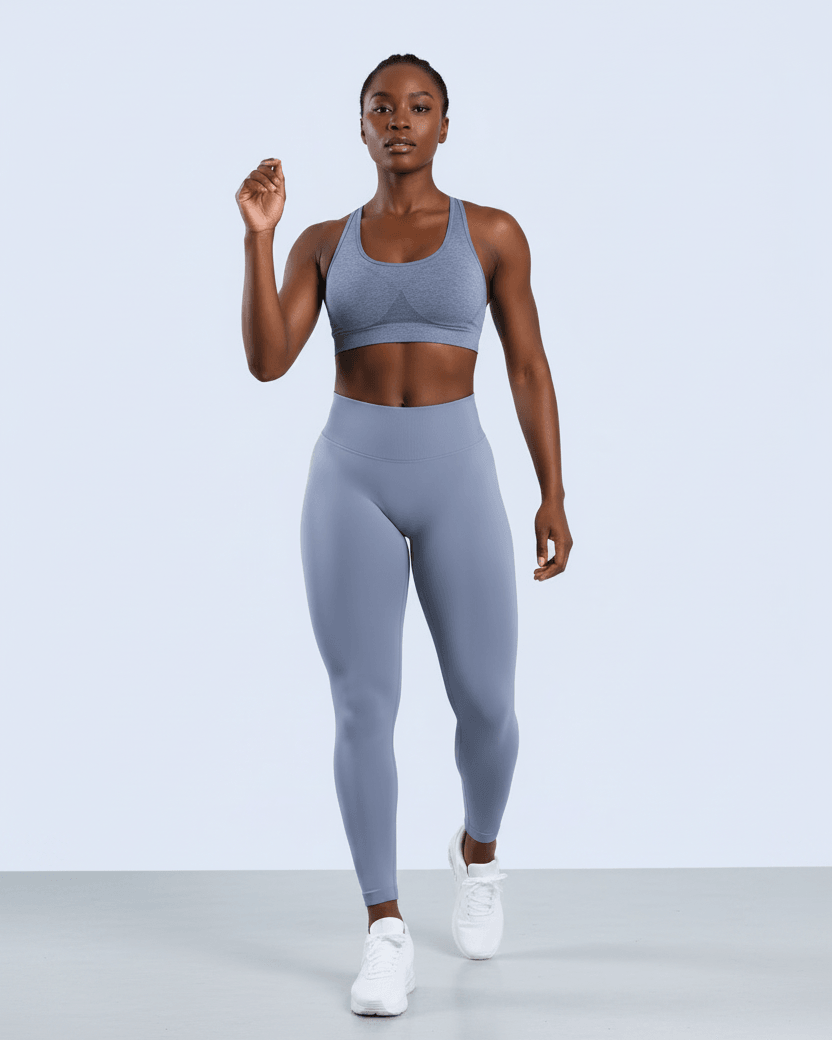 A woman modeling a matching athletic set consisting of a light blue-gray sports bra and high-waisted leggings, paired with white sneakers, against a clean light gray studio background.