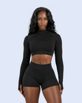 A woman modeling a black long-sleeve crop top with a half-zip neckline and matching black high-waisted athletic shorts against a light blue studio background.