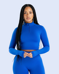 A woman with long black braids wearing a bright royal blue long-sleeve crop top with a mock neck and matching high-waisted leggings, posed against a light gray studio background.